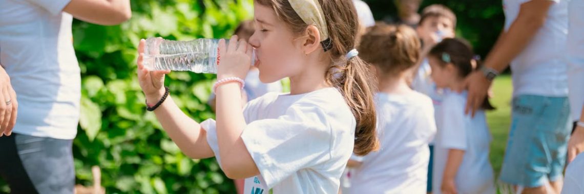 Young girl drinking water during sunny outdoor activity with friends in a park setting.